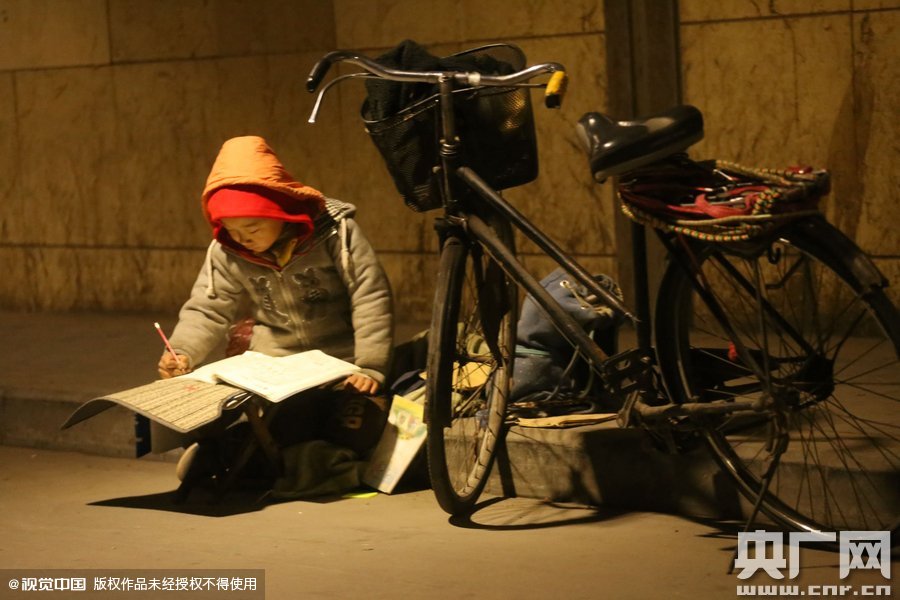 鄭州中原路京廣鐵路大橋下涵洞的馬路牙子邊，一位戴著紅色辣椒帽的小男孩趴在一個(gè)小馬扎上，上面鋪著一張車墊，在微弱的燈光下寫作業(yè)